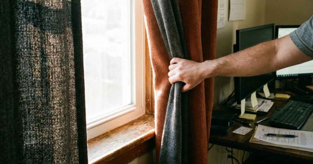 A Home Energy Expert Reveals Why Remote Work Is Quietly Doubling Your Electric Bill 4 Hand closing heavy curtains on a sunny window next to a desk to block heat.